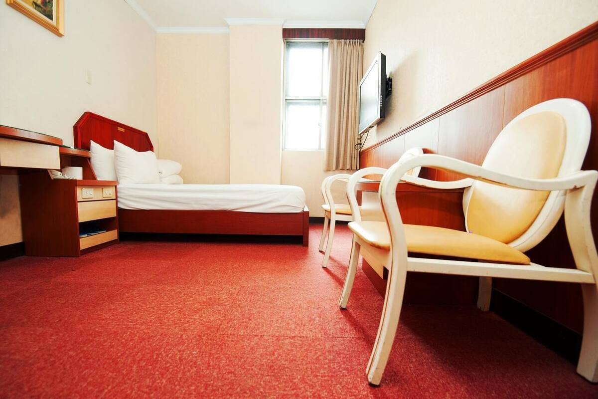 A hotel room with a bed, a desk with a telephone, a chair, a television mounted on the wall, and a window with curtains.