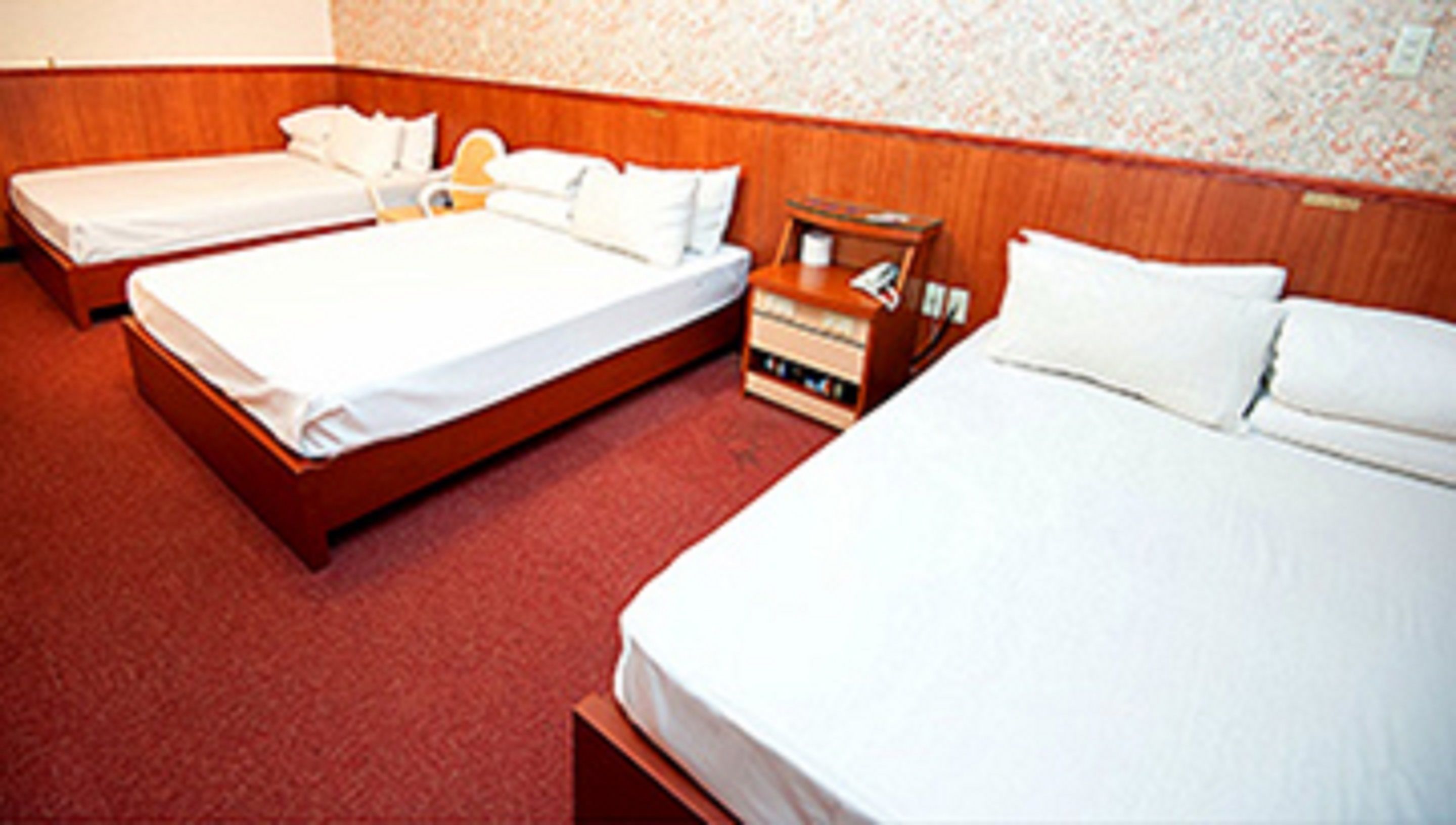 Una habitación de hotel con dos camas individuales, un cabecero de madera, una mesita de noche pequeña con una lámpara y suelo alfombrado.