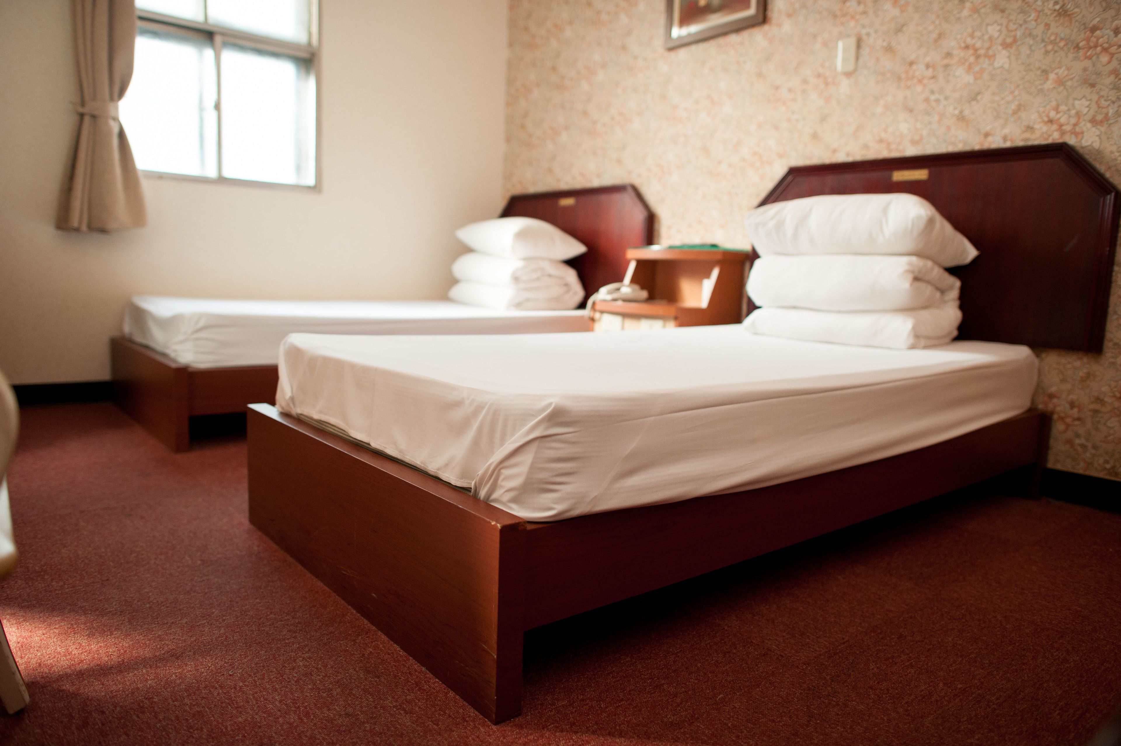 A hotel room with two single beds, a wooden headboard, a small bedside table, and a window with curtains.