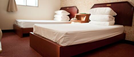 A hotel room with two single beds, a wooden headboard, a small bedside table, and a window with curtains.