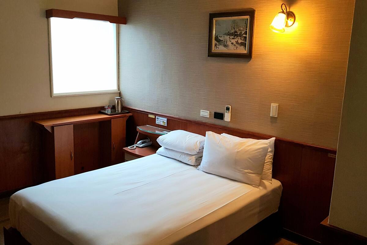 A hotel room with a bed, a nightstand, a desk, a window, and a wall-mounted light fixture.