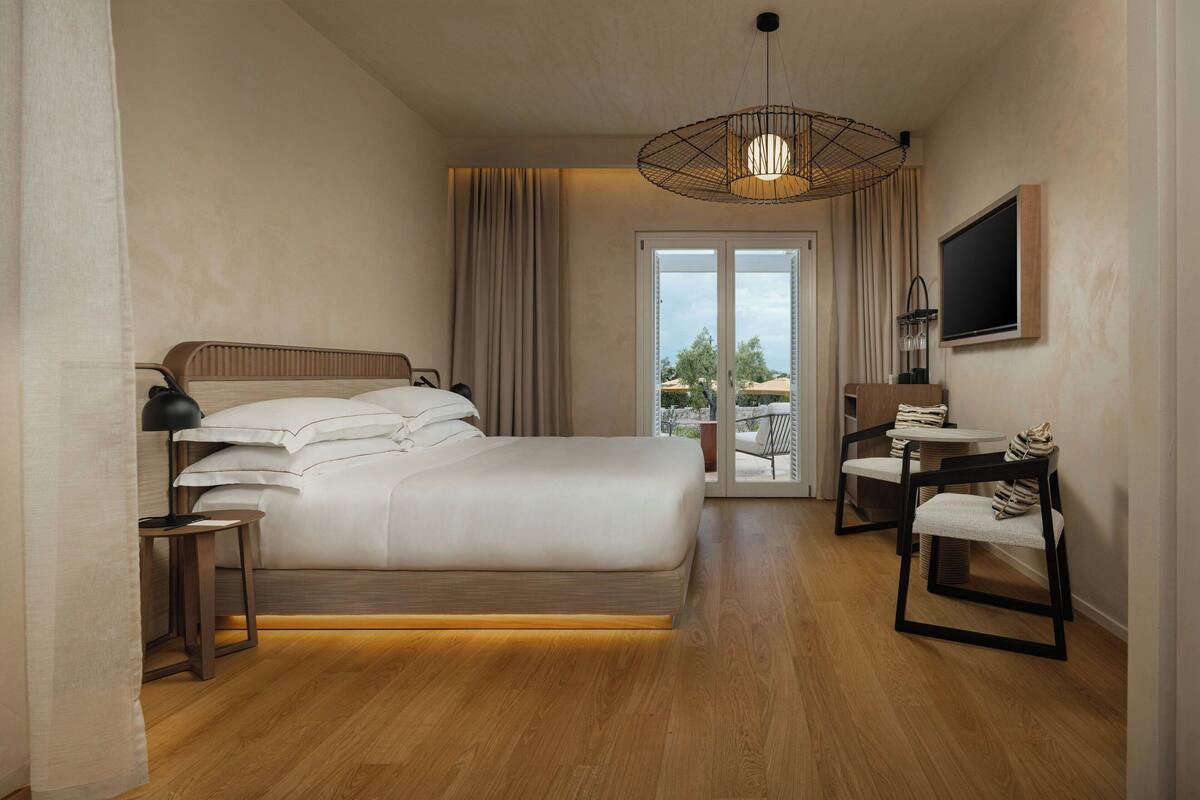 Ein modernes Hotelzimmer mit einem großen Bett, einem Nachttisch mit Lampe, einem an der Wand befestigten Fernseher und einem Balkon mit Sitzgelegenheit.
