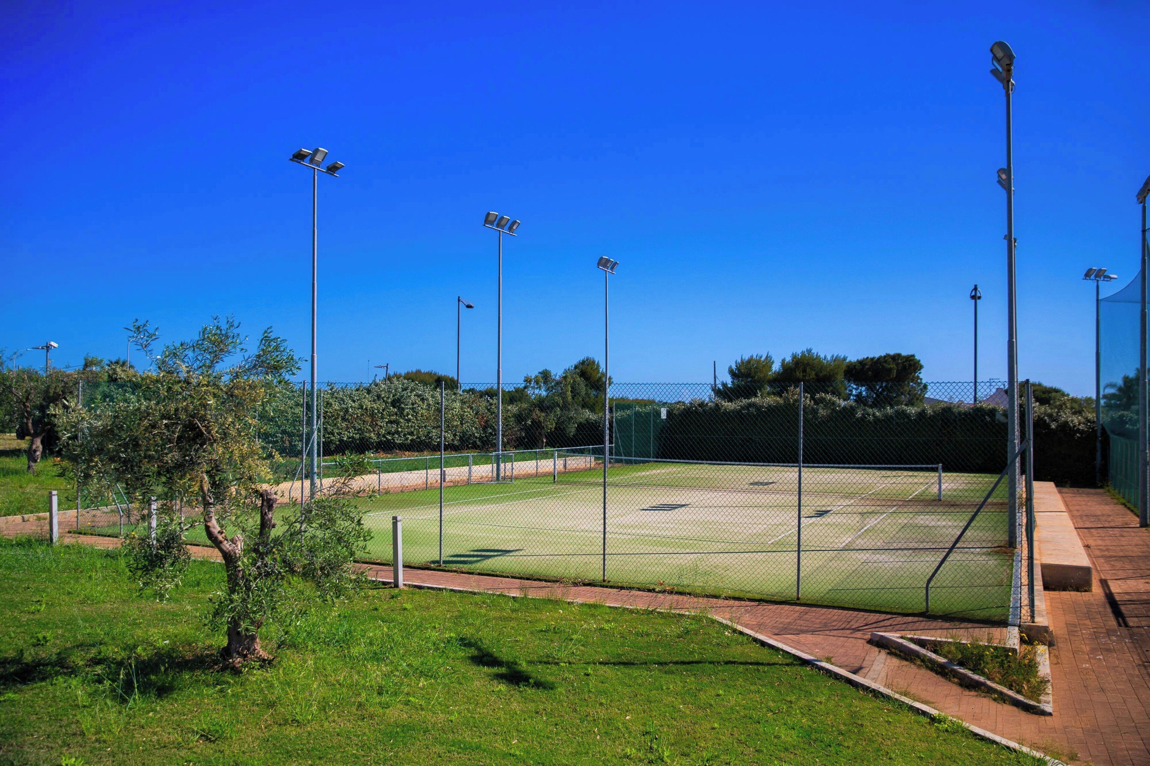 A tennis court with surrounding fencing, trees, and street lamps.