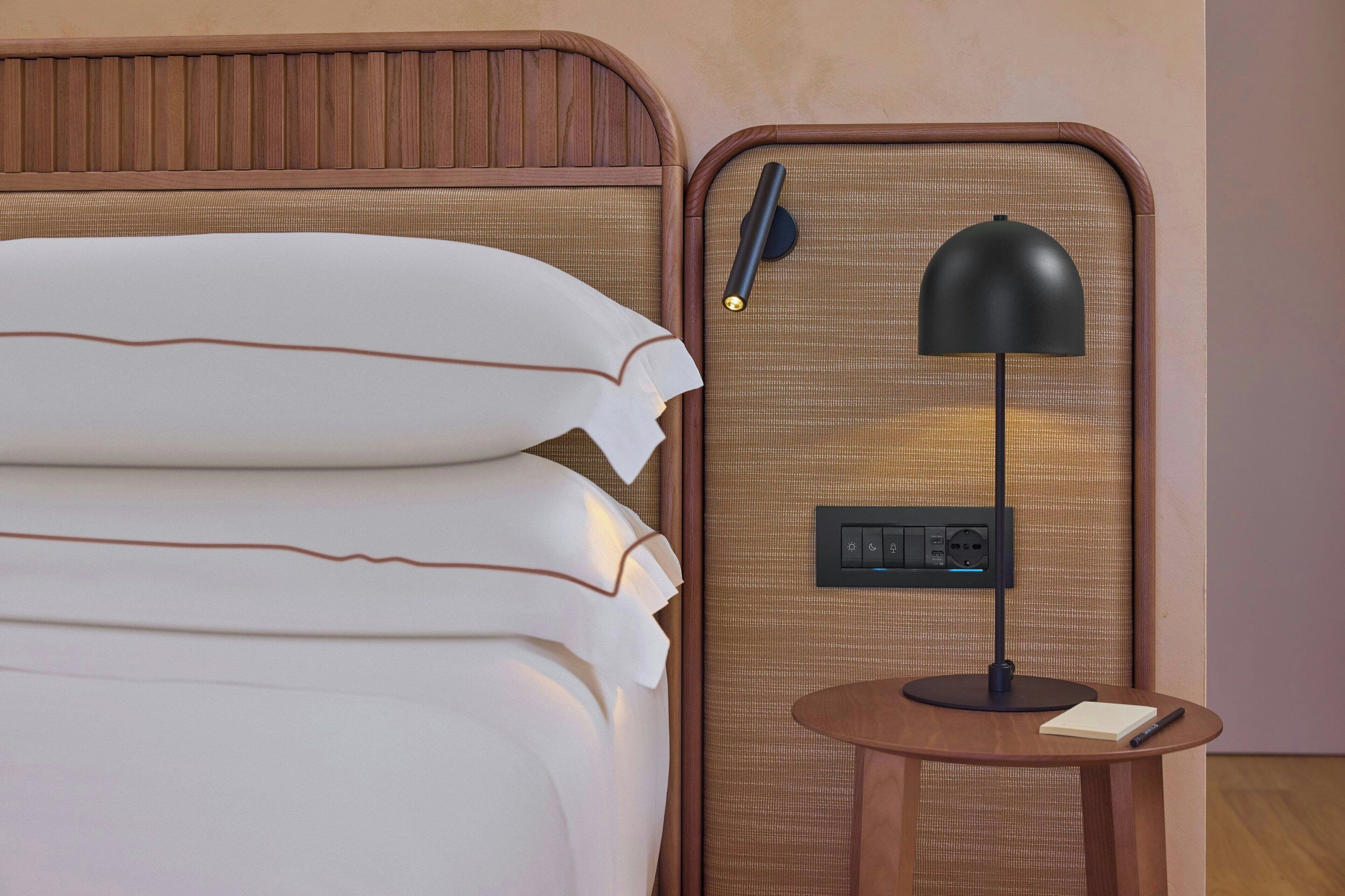 Ein ordentlich bezogenes Bett mit weißen Leinen, ein kleiner Nachttisch mit einer schwarzen Lampe und einem Notizblock.