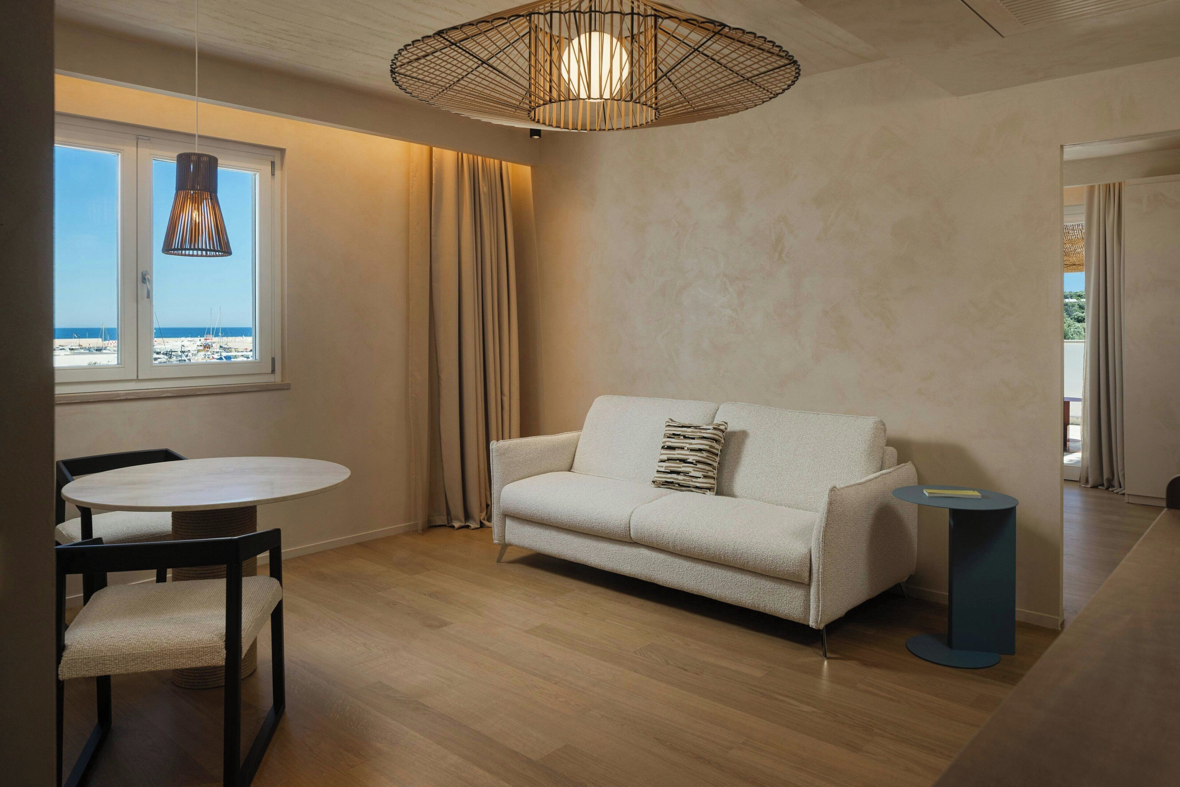 Un salon moderne avec un canapé, une table ronde et des chaises. La fenêtre offre une vue sur la plage.
