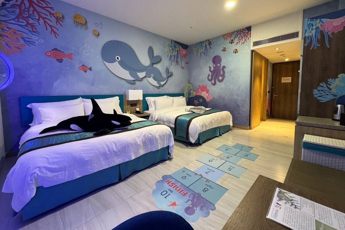 Une chambre d’hôtel avec deux lits, une grande fresque représentant des créatures marines sur le mur et un jeu de marelle peint sur le sol.