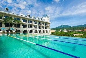 A large swimming pool with clear blue water, surrounded by a white building with multiple balconies and a mountain view in the background.