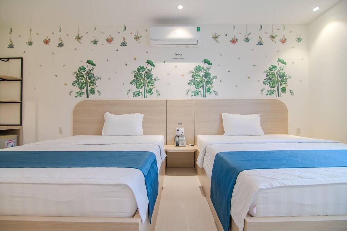 A hotel room with two beds, a wooden headboard, a small bedside table with a phone, and a wall decorated with hanging plants and flowers.