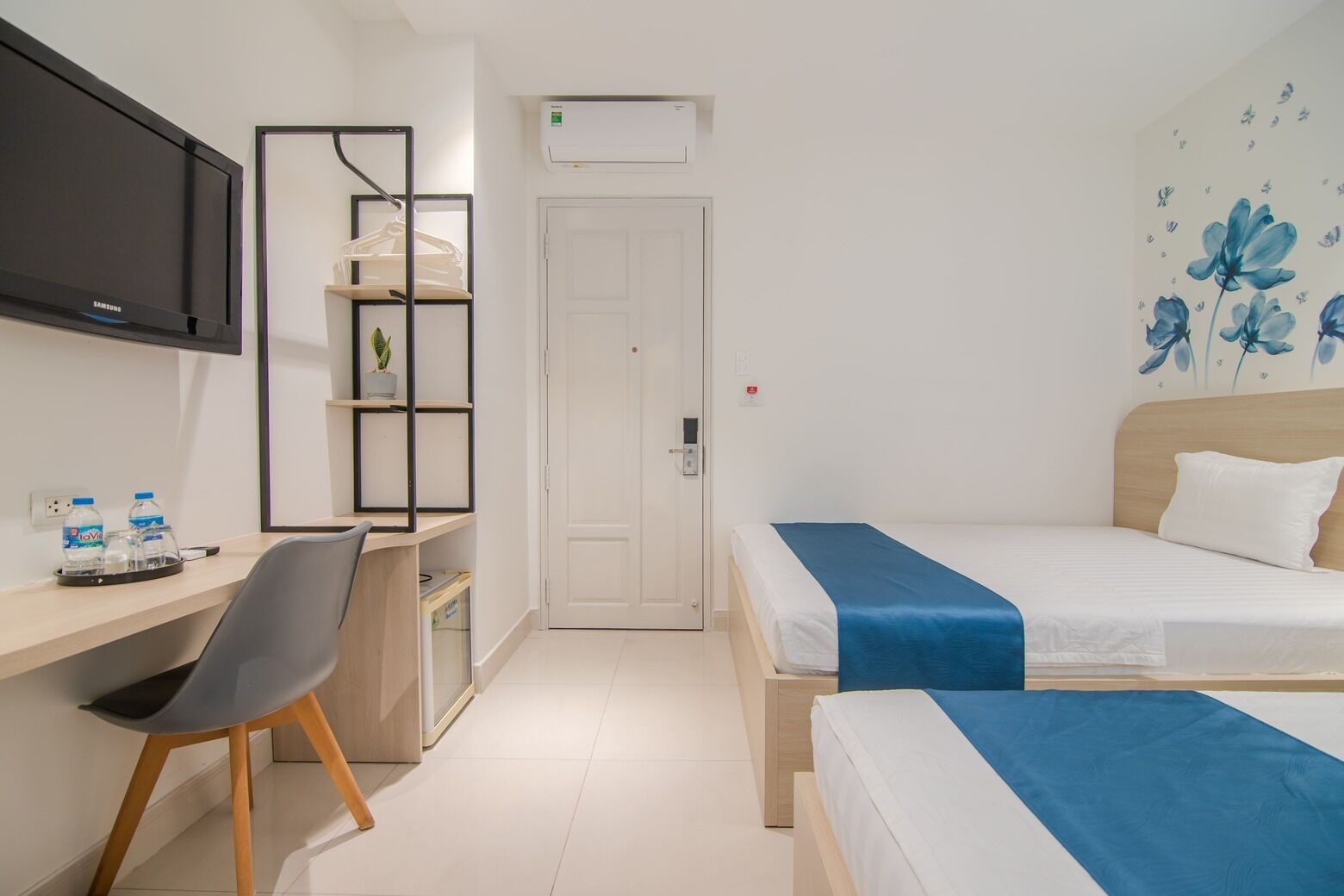 A hotel room with a bed, a desk with a chair, a TV, and a wall with a floral design.