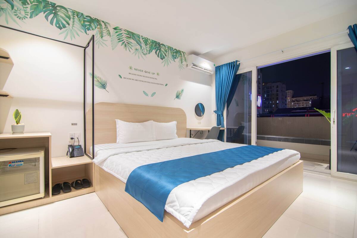 A modern hotel room with a large bed, a bedside table with a lamp, a small desk with a chair, a potted plant, and a view of the city through a large window.
