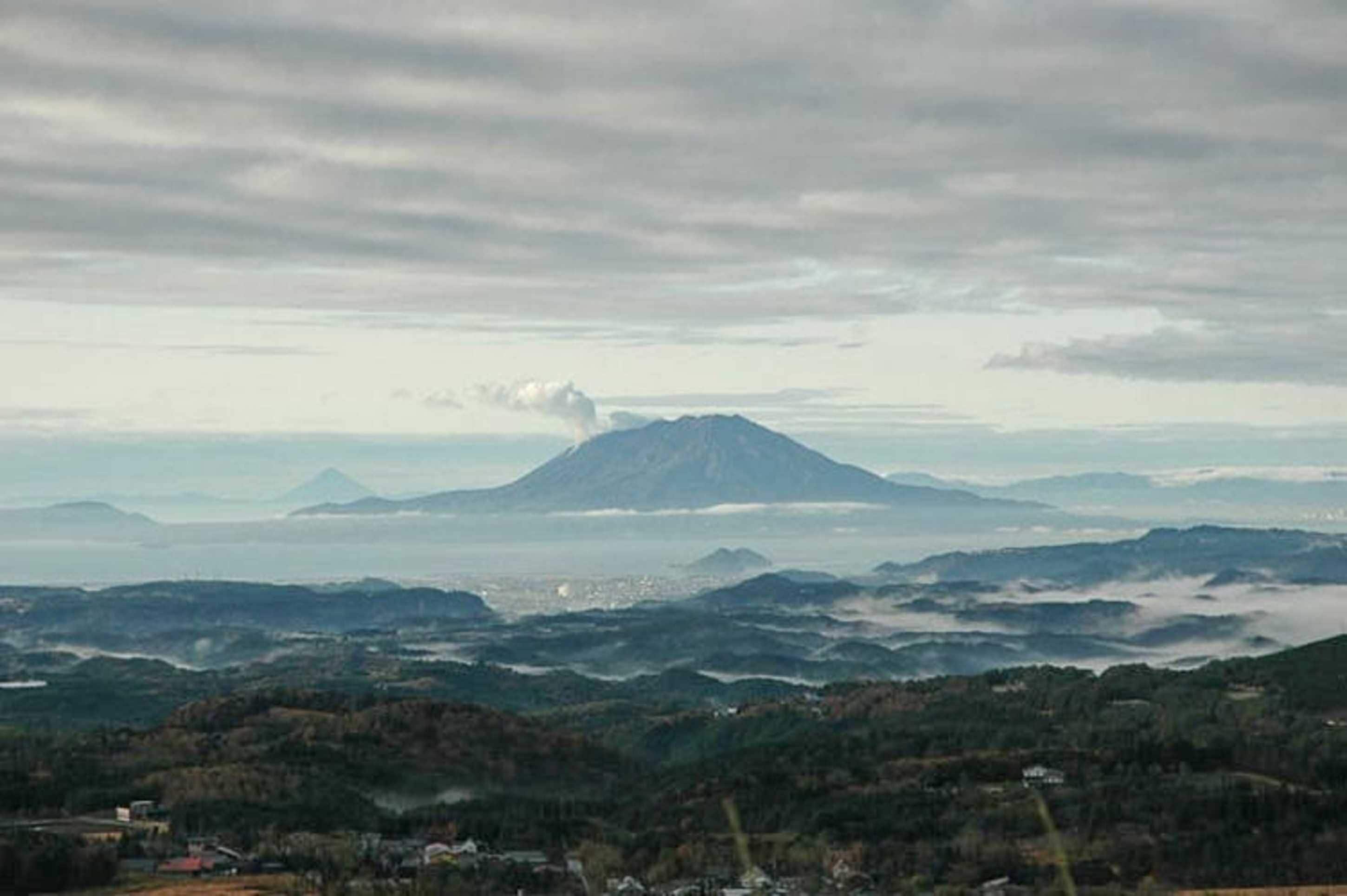 A mountain with a visible smoke plume, surrounded by a valley with scattered buildings and dense forest.