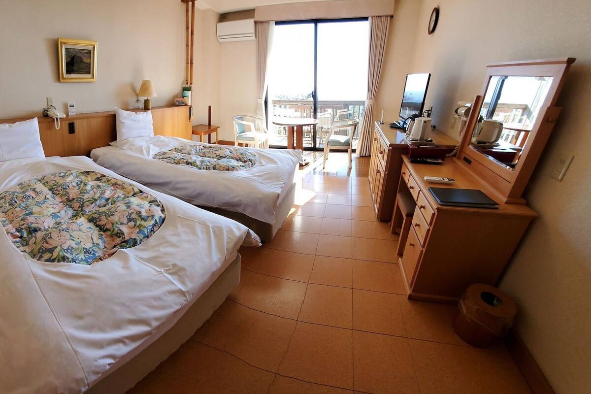 A hotel room with two beds, a desk, a chair, a TV, and a balcony with a view.