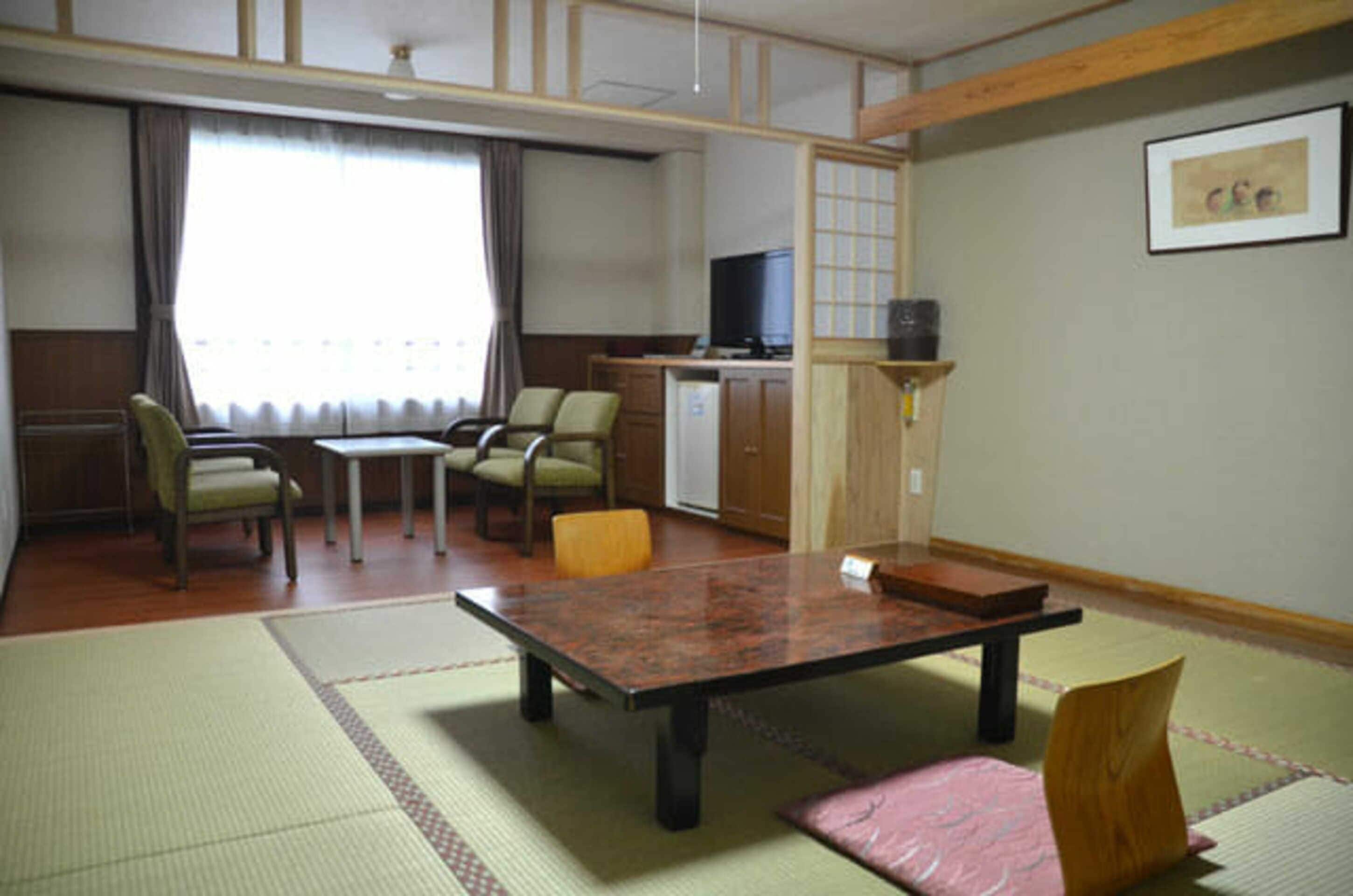 A traditional Japanese room with tatami flooring, a low table, chairs, a television, and a small kitchen area.