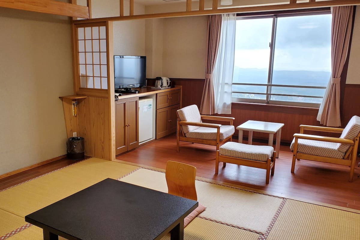 A room with tatami flooring, a black table, wooden chairs, a TV, a coffee maker, and a view of the sea through a window.