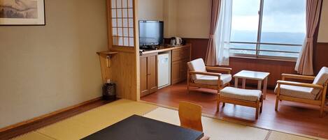 Una habitación con suelo de tatami, una mesa negra, sillas de madera, un televisor, una cafetera y vista al mar a través de una ventana.