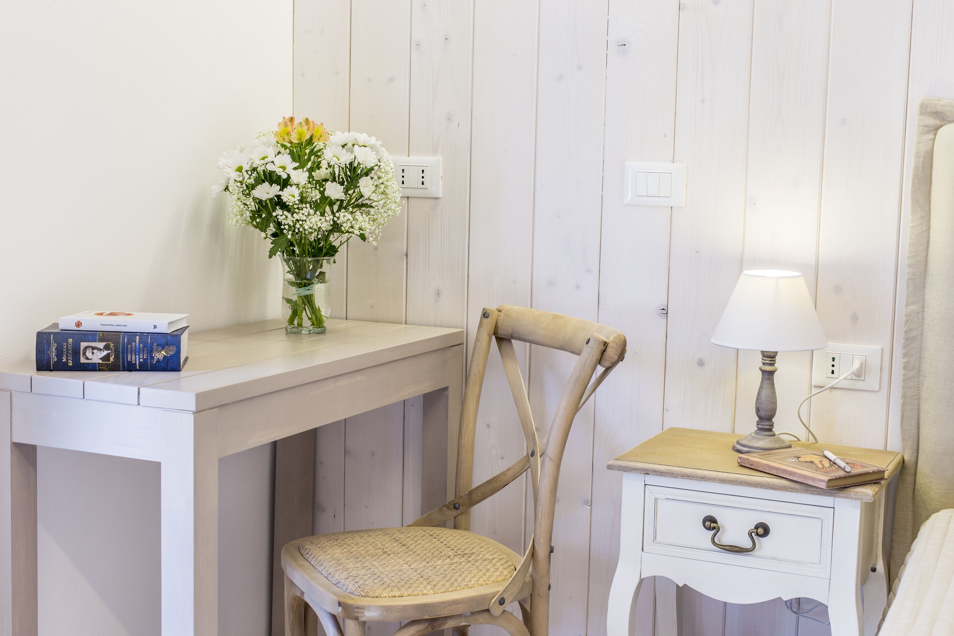 A room with a wooden desk, a chair, a bedside table with a lamp, and a vase with flowers.