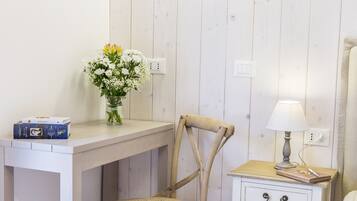 A room with a wooden desk, a chair, a bedside table with a lamp, and a vase with flowers.