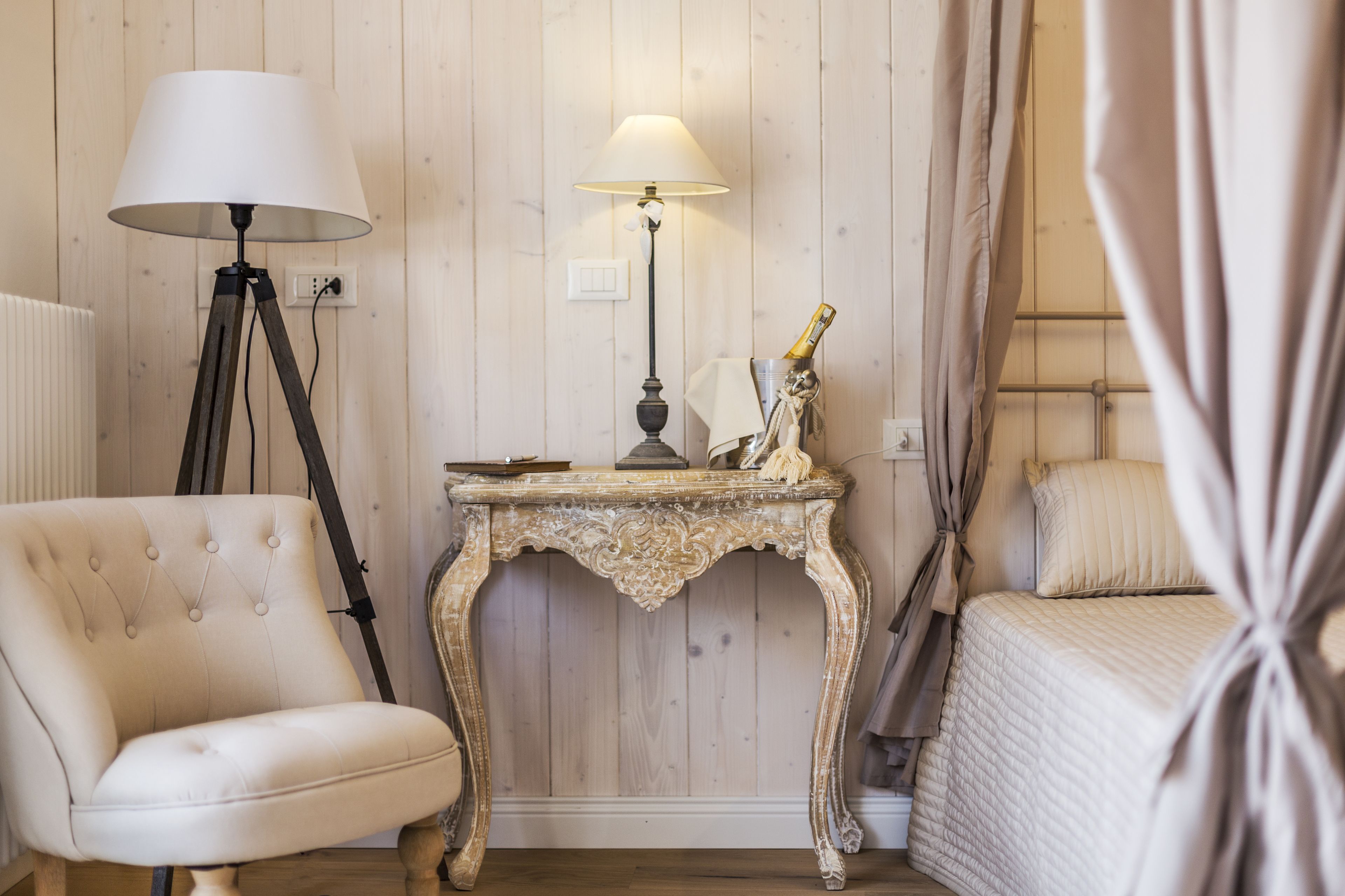 A room with a wooden paneled wall, a white upholstered armchair, a small ornate table with a lamp, and a bed with curtains.
