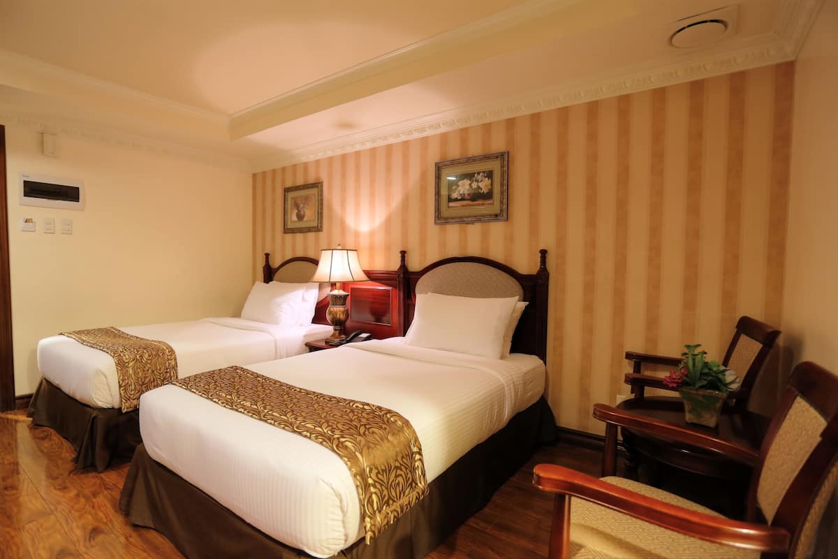 Habitación de hotel con dos camas, una mesita de noche, una lámpara, una silla y una pequeña mesa con una planta.