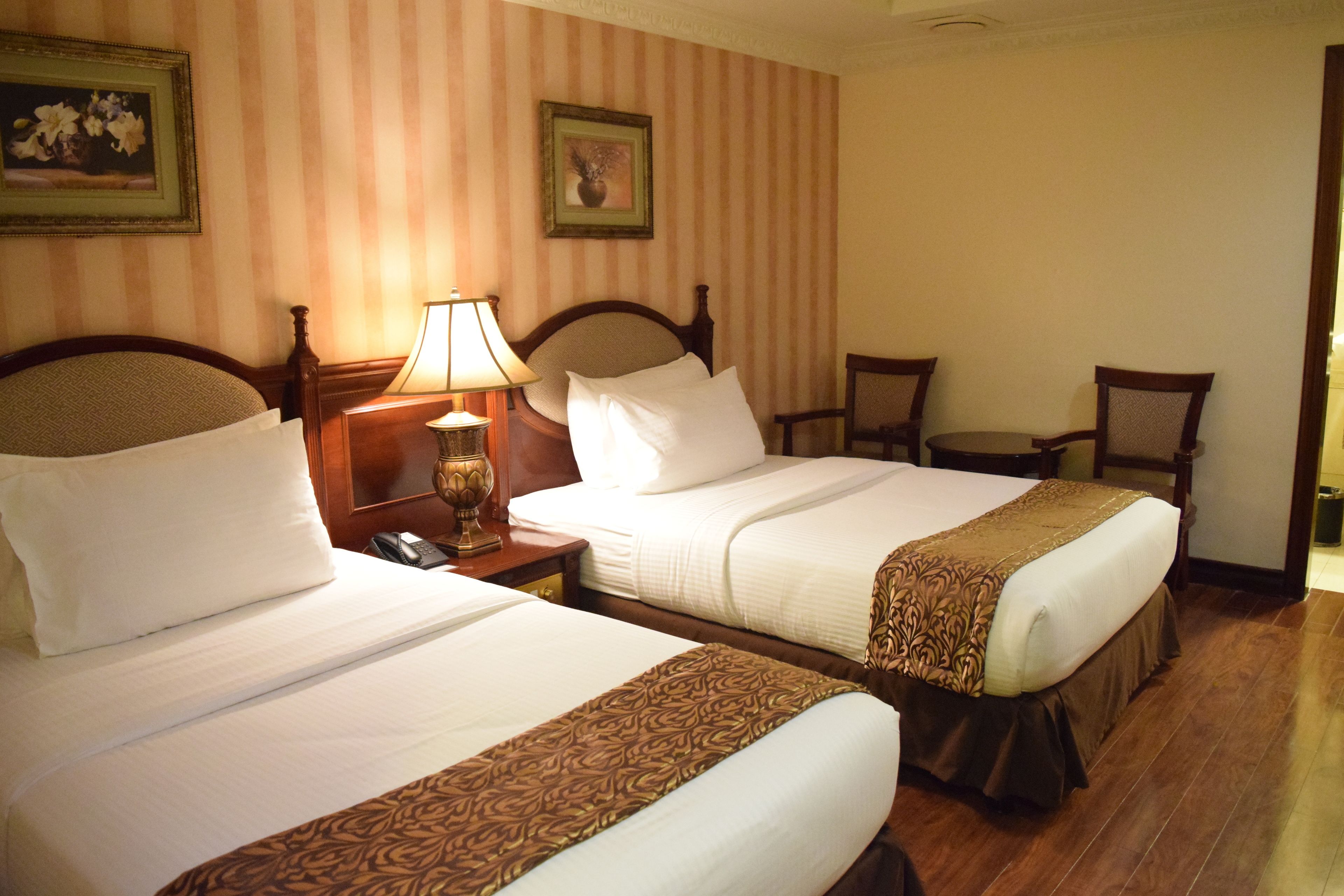 A hotel room with two beds, a nightstand with a lamp, a chair, and a small table.