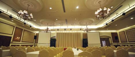 Un salón de banquetes con filas de mesas y sillas, candelabros ornamentales y un escenario con cortinas.
