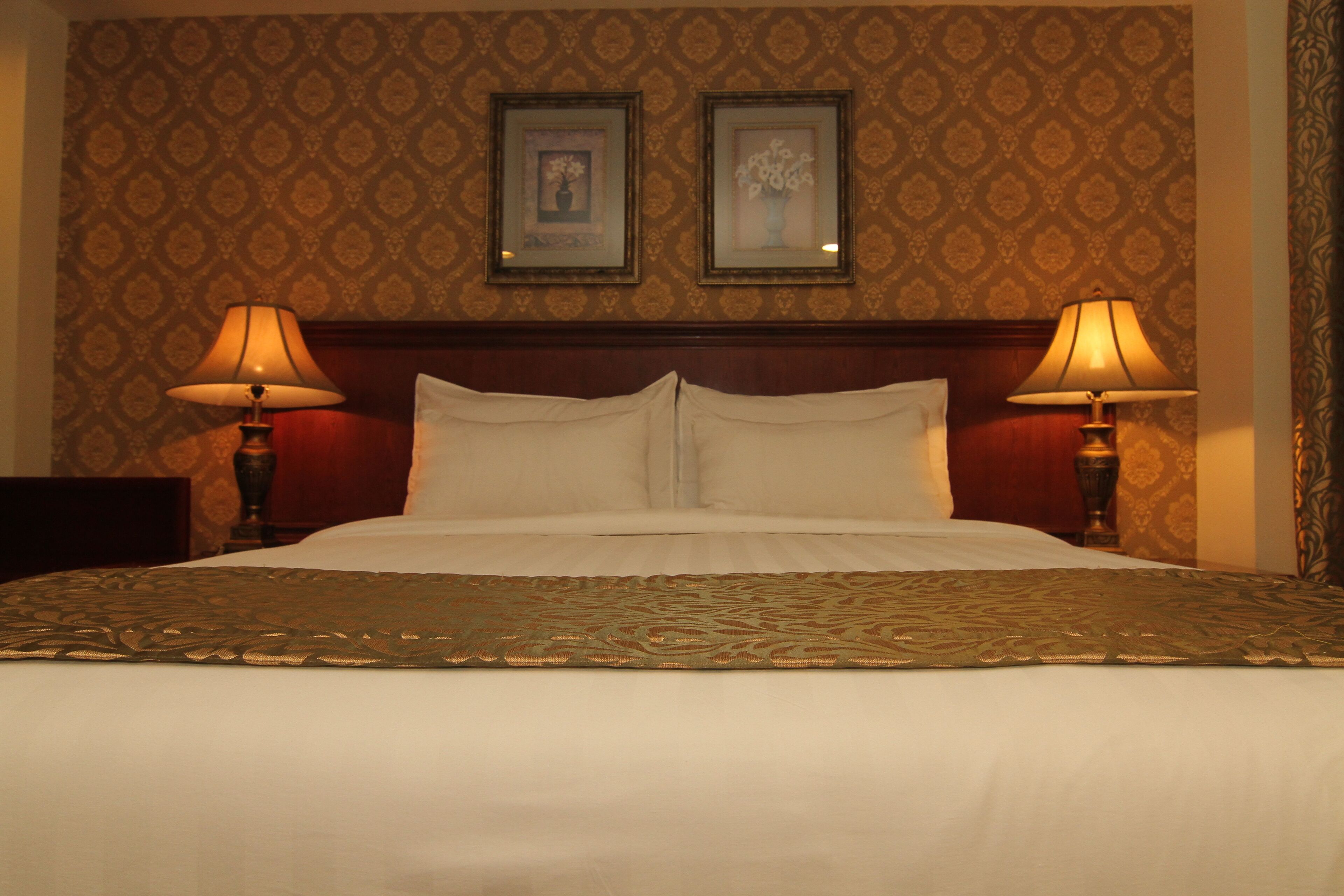 A hotel room with a large bed, two bedside lamps, a headboard, and two framed pictures on the wall.