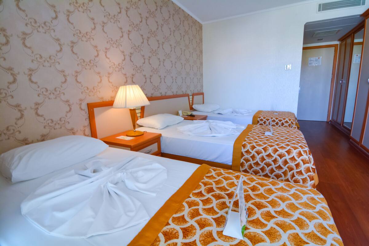 Ein Hotelzimmer mit drei Betten, jedes mit weißer Bettwäsche und einer gemusterten Tagesdecke. Ein Nachttisch mit Lampe und Telefon ist vorhanden. Die Wände sind mit strukturiertem Wandpapier versehen und der Boden ist aus Holz.