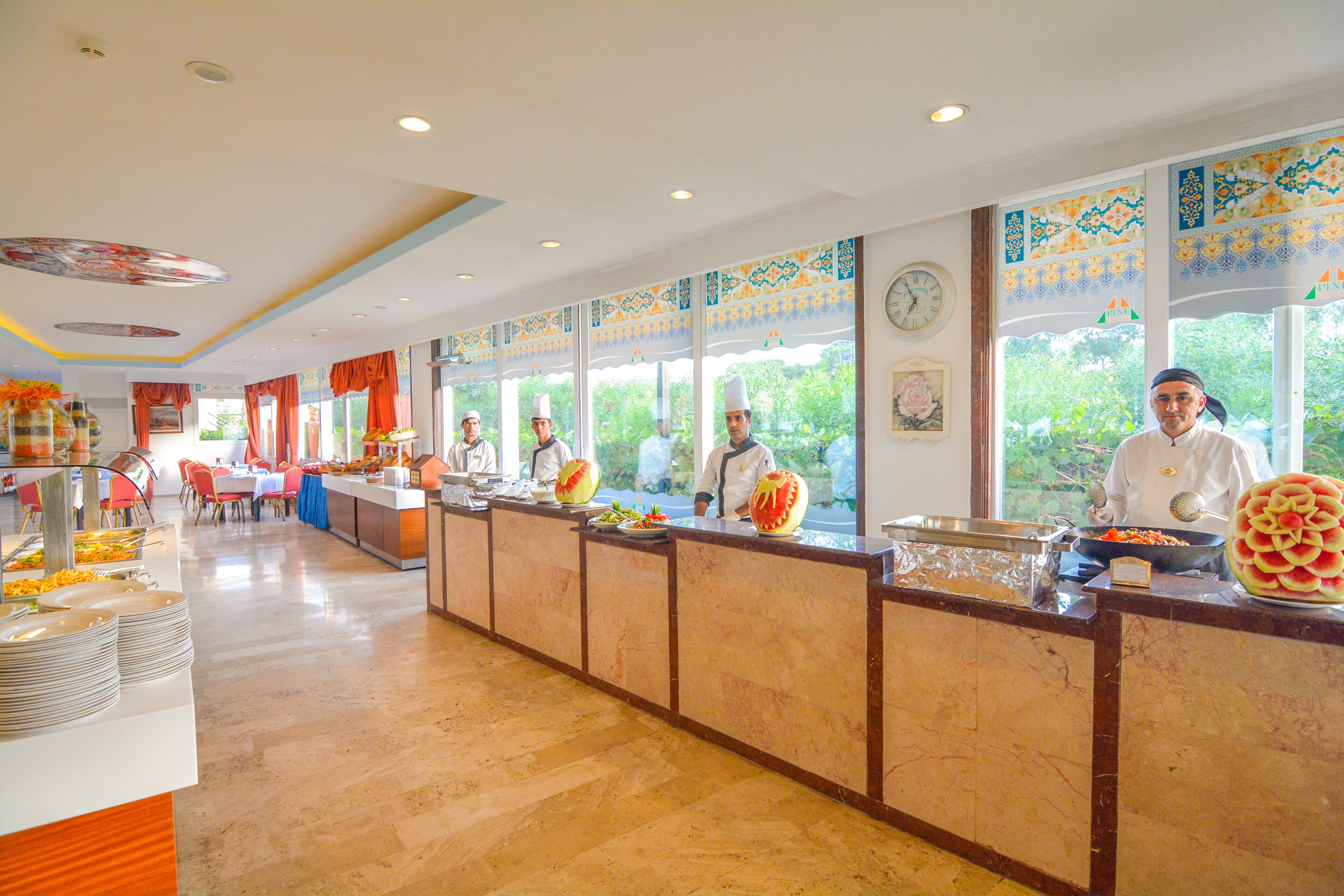 A buffet area with multiple serving stations, a clock on the wall, and a view of greenery outside.