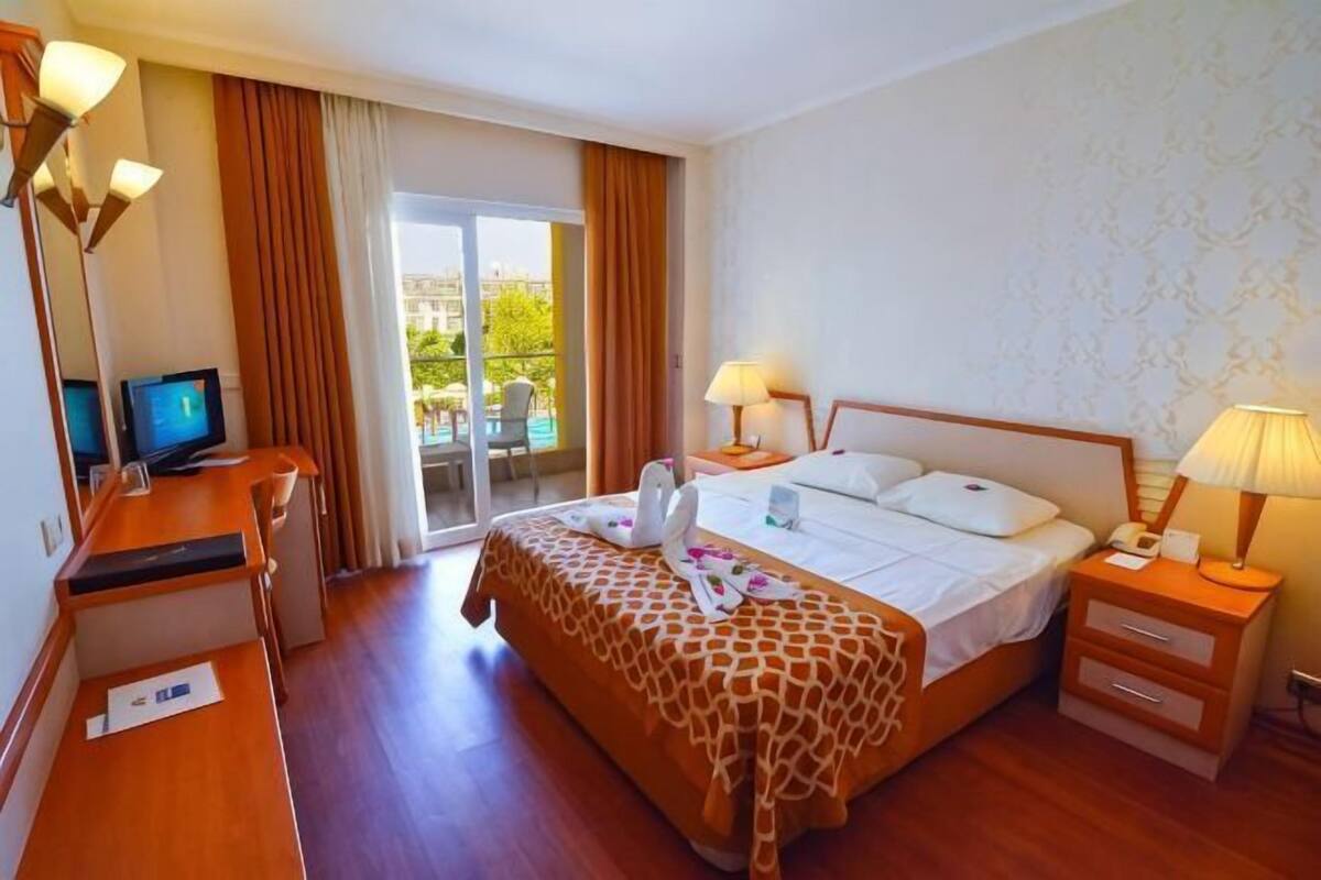 A hotel room with a bed, bedside tables, a desk with a computer, and a view of the outdoors.