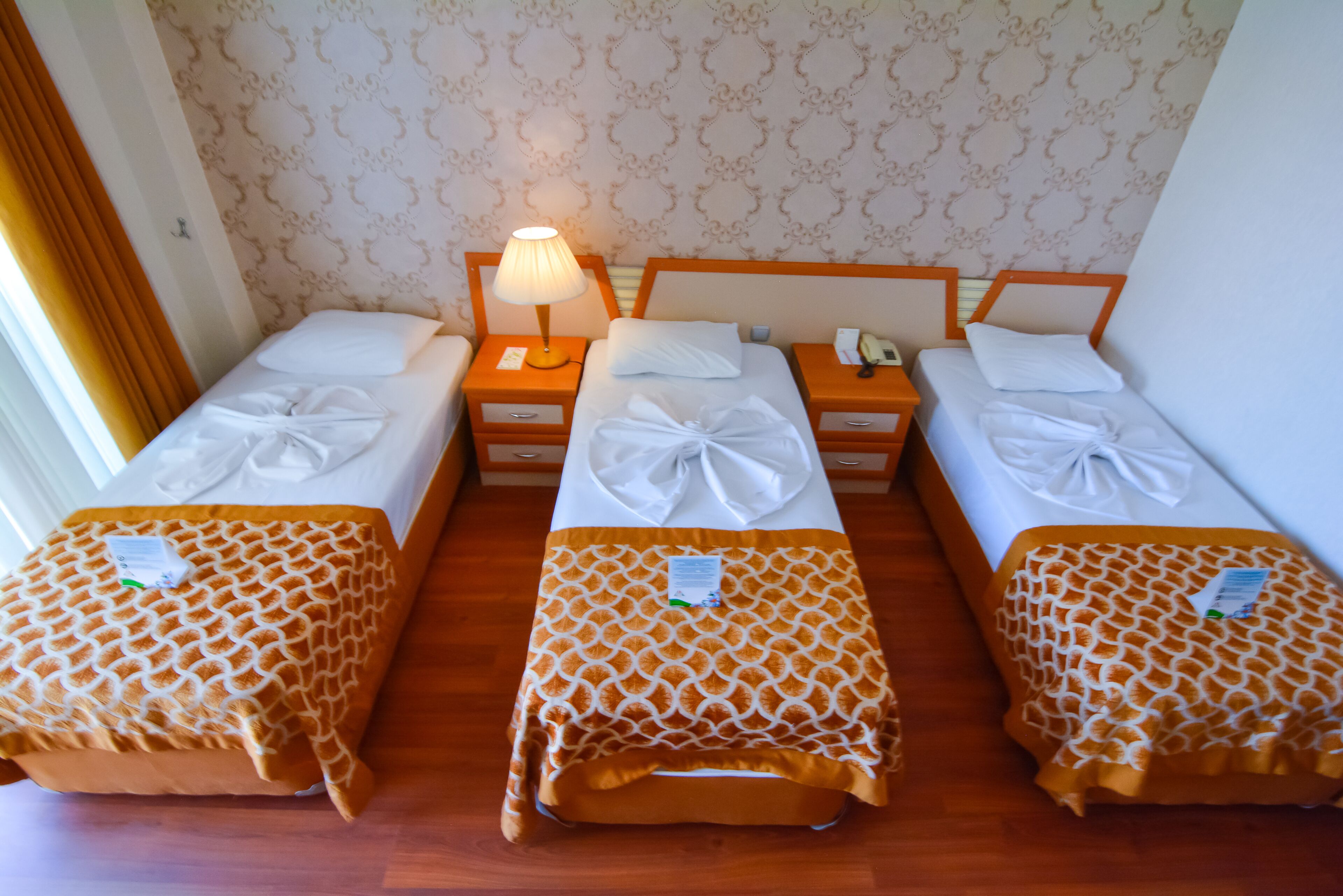 Habitación de hotel con tres camas individuales, cada una con una colcha estampada y almohada bien ordenada. Hay una mesita de noche con lámpara entre las camas, y un teléfono en la mesita de noche derecha.