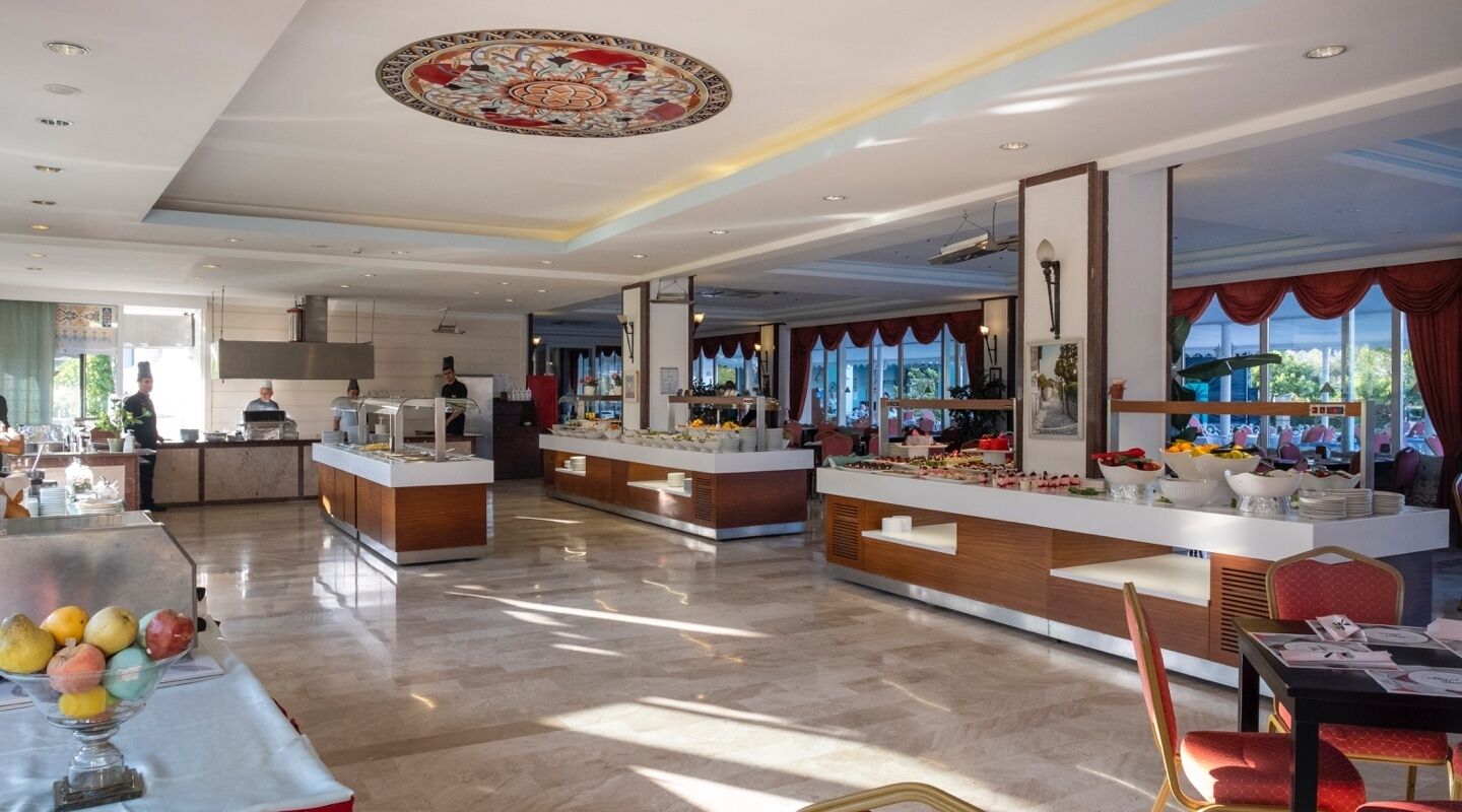 Un moderno vestíbulo de hotel con un mostrador de recepción central, una zona de buffet y un comedor con vistas al exterior.