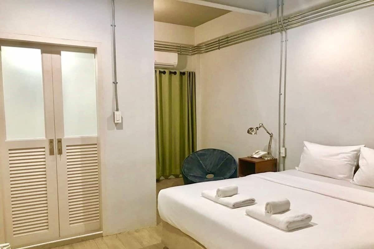 Quarto de hotel com uma cama, mesa de cabeceira, ventilador, cortina verde e guarda-roupa.