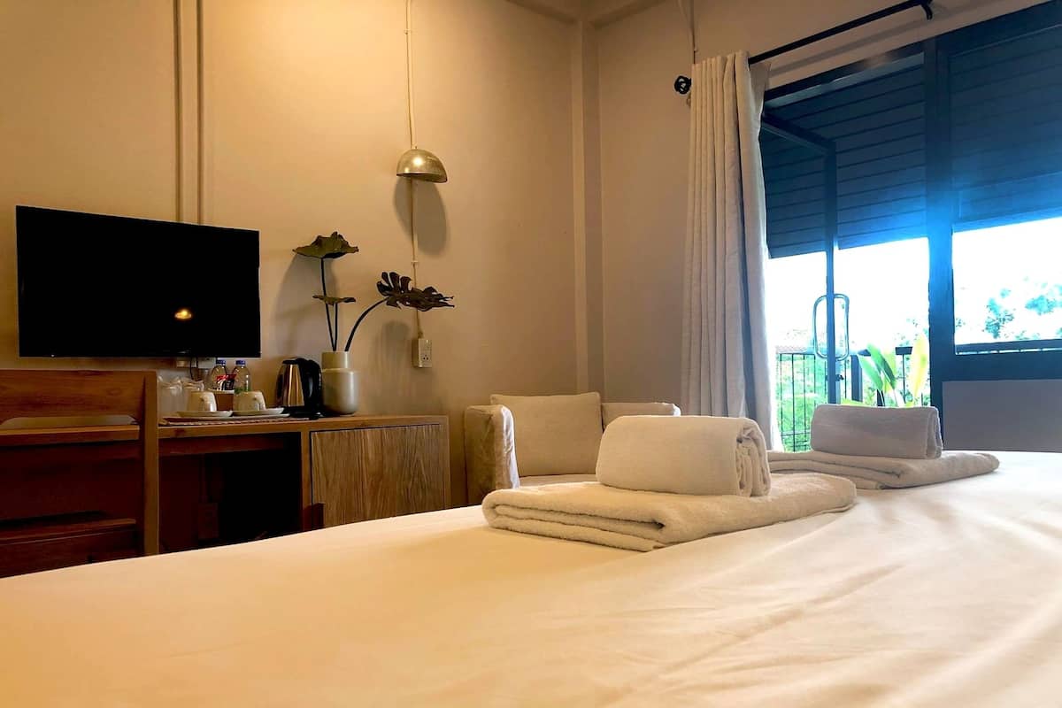 Quarto de hotel com cama, escrivaninha com televisão, cadeira, janela com cortinas e vista para o verde lá fora.