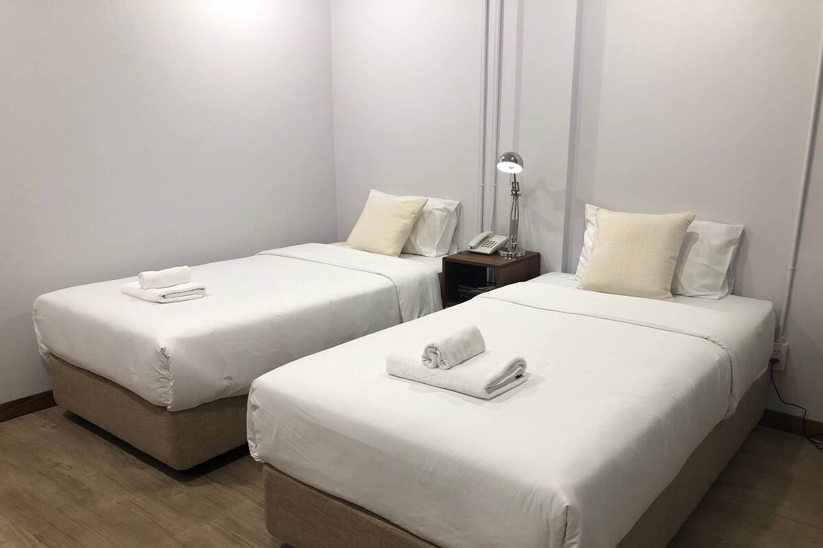 Quarto de hotel com duas camas de solteiro, cada uma com um edredom branco e uma toalha enrolada. Há uma mesinha de cabeceira com uma luminária e um telefone.