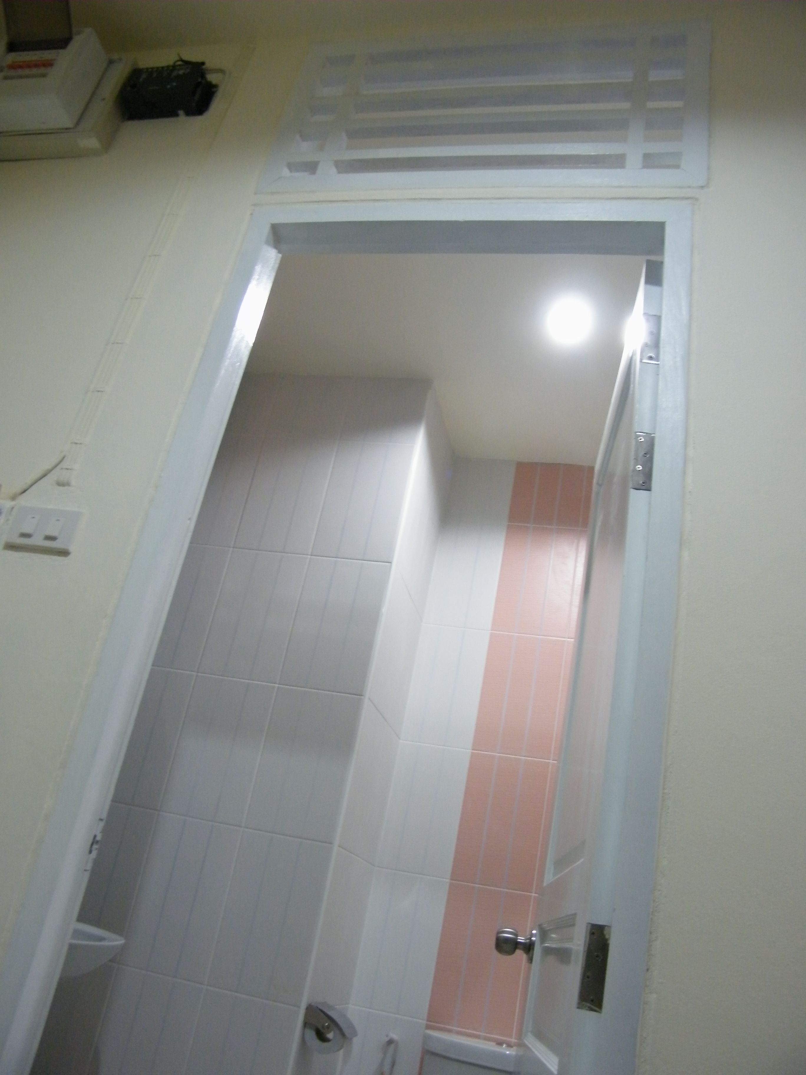 A narrow bathroom with white and pink tiles, a white door, and a light fixture above the door.