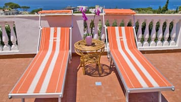 Two striped lounge chairs with a small round table and a purple flower between them, overlooking a coastal view.