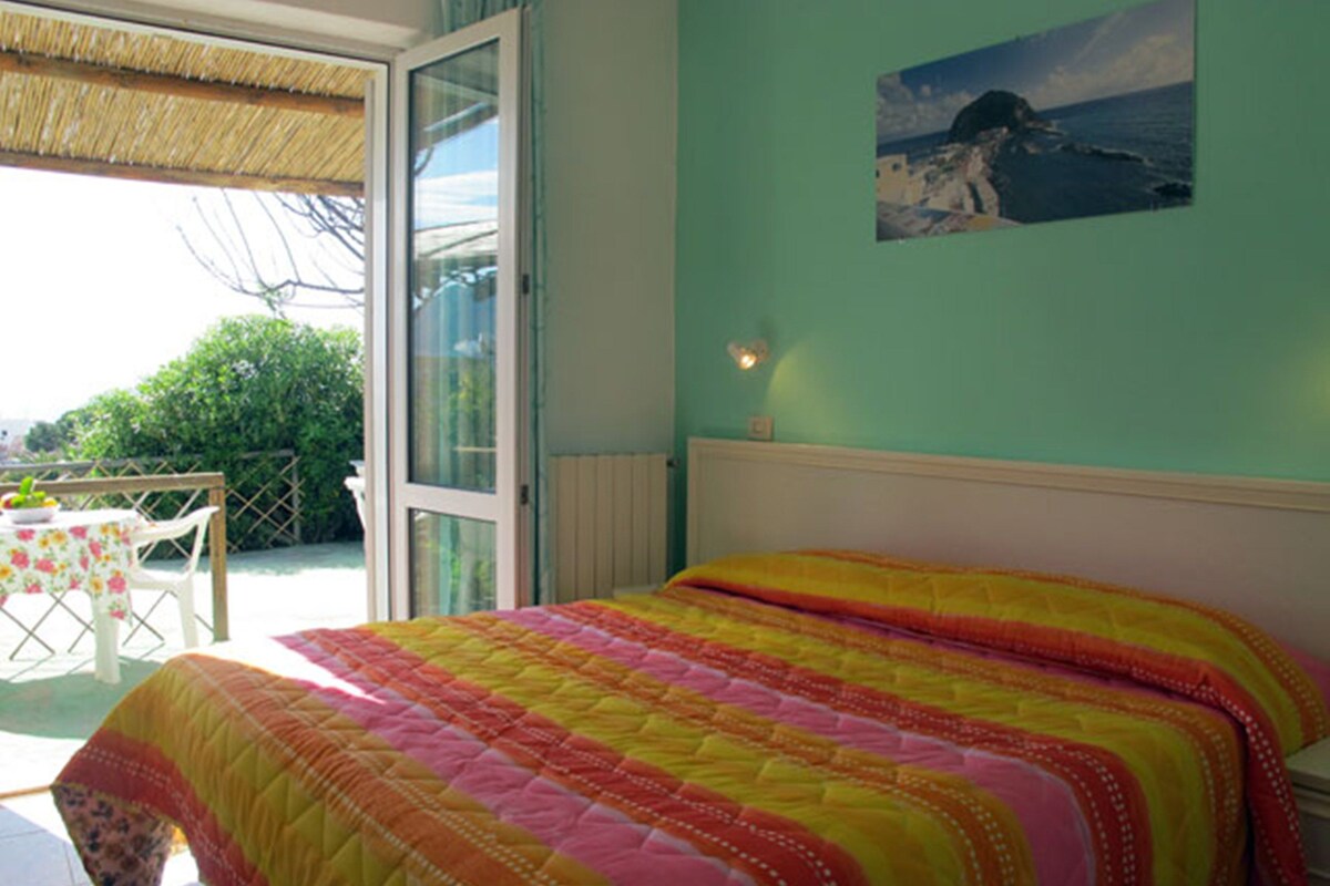 A bedroom with a colorful quilted bedspread, a view of the outdoors through a glass door, and a painting on the wall.