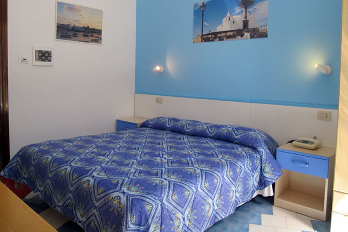 A bedroom with a bed, a nightstand, a wall-mounted lamp, and two framed pictures.