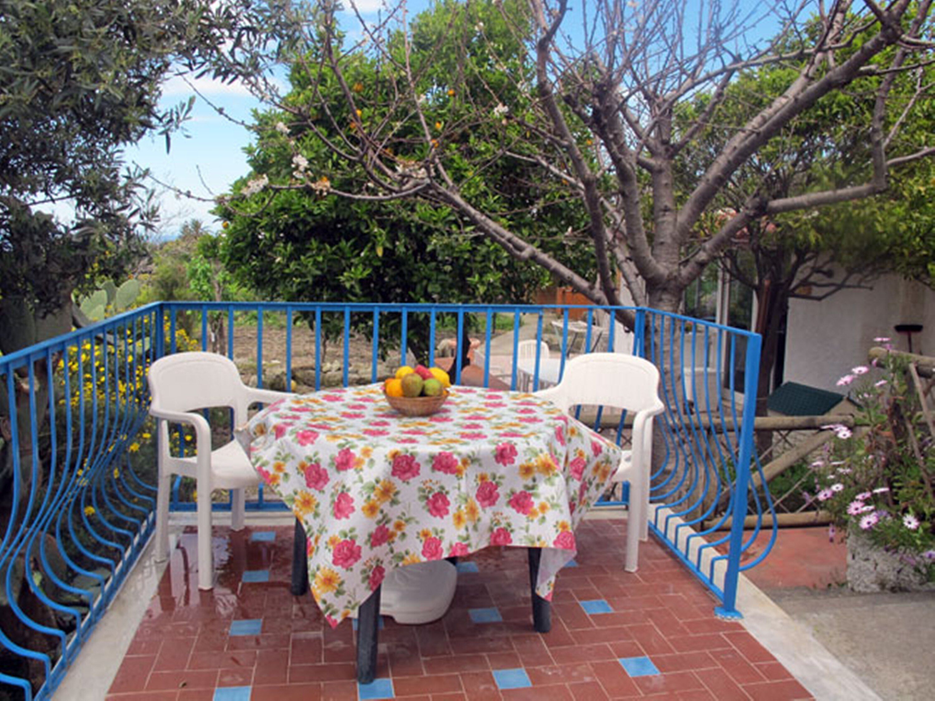 A table with a floral tablecloth, two white chairs, and a basket of fruit.