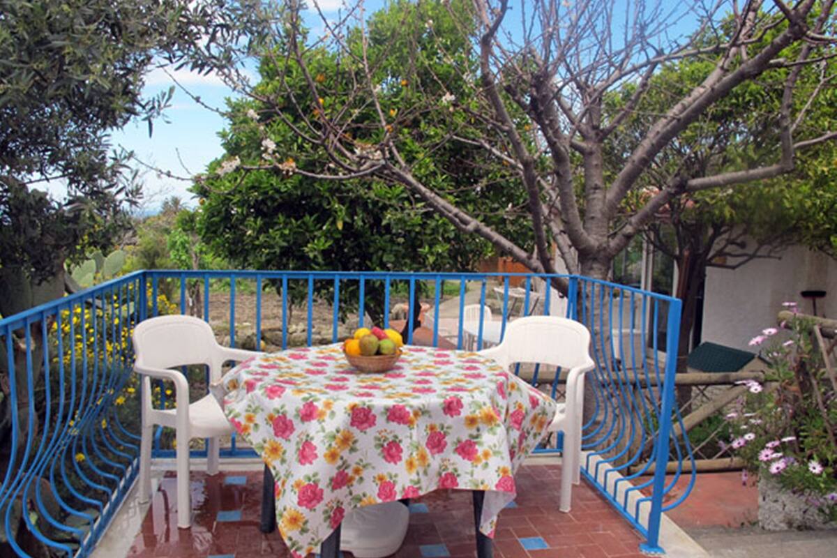 A table with a floral tablecloth, two white chairs, and a basket of fruit.