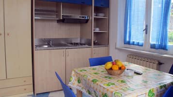 Une cuisine compacte équipée d’un évier, d’une cuisinière et de meubles bleus. Une table avec un panier de fruits et des chaises se trouve devant une fenêtre ornée de rideaux bleus.