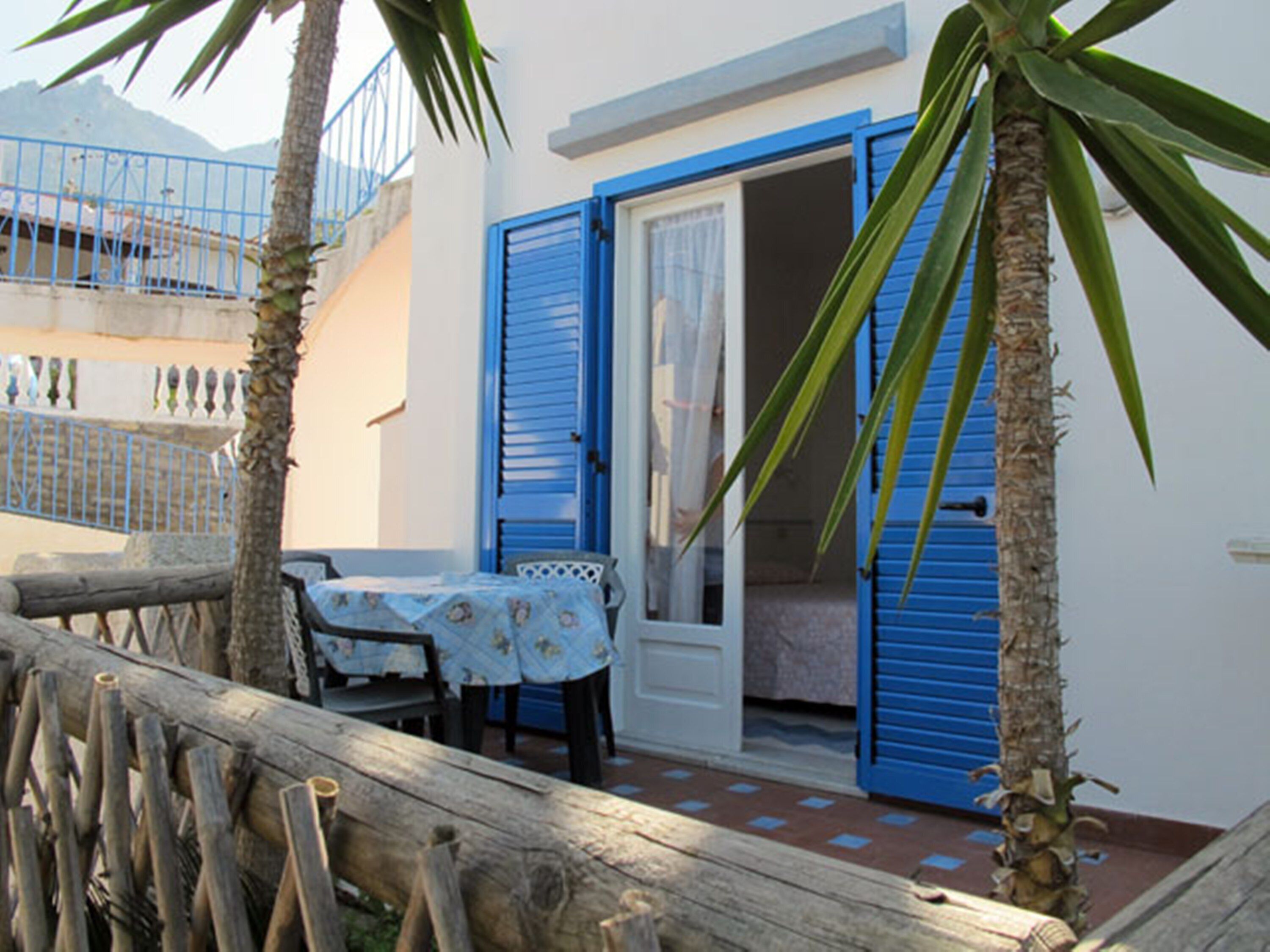A small outdoor seating area with a table and chairs, a wooden fence, and blue shutters on the building.