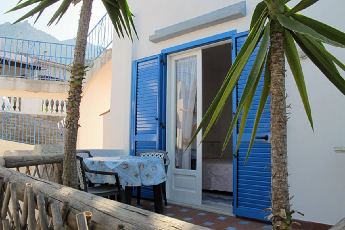 A small outdoor seating area with a table and chairs, a wooden fence, and blue shutters on the building.