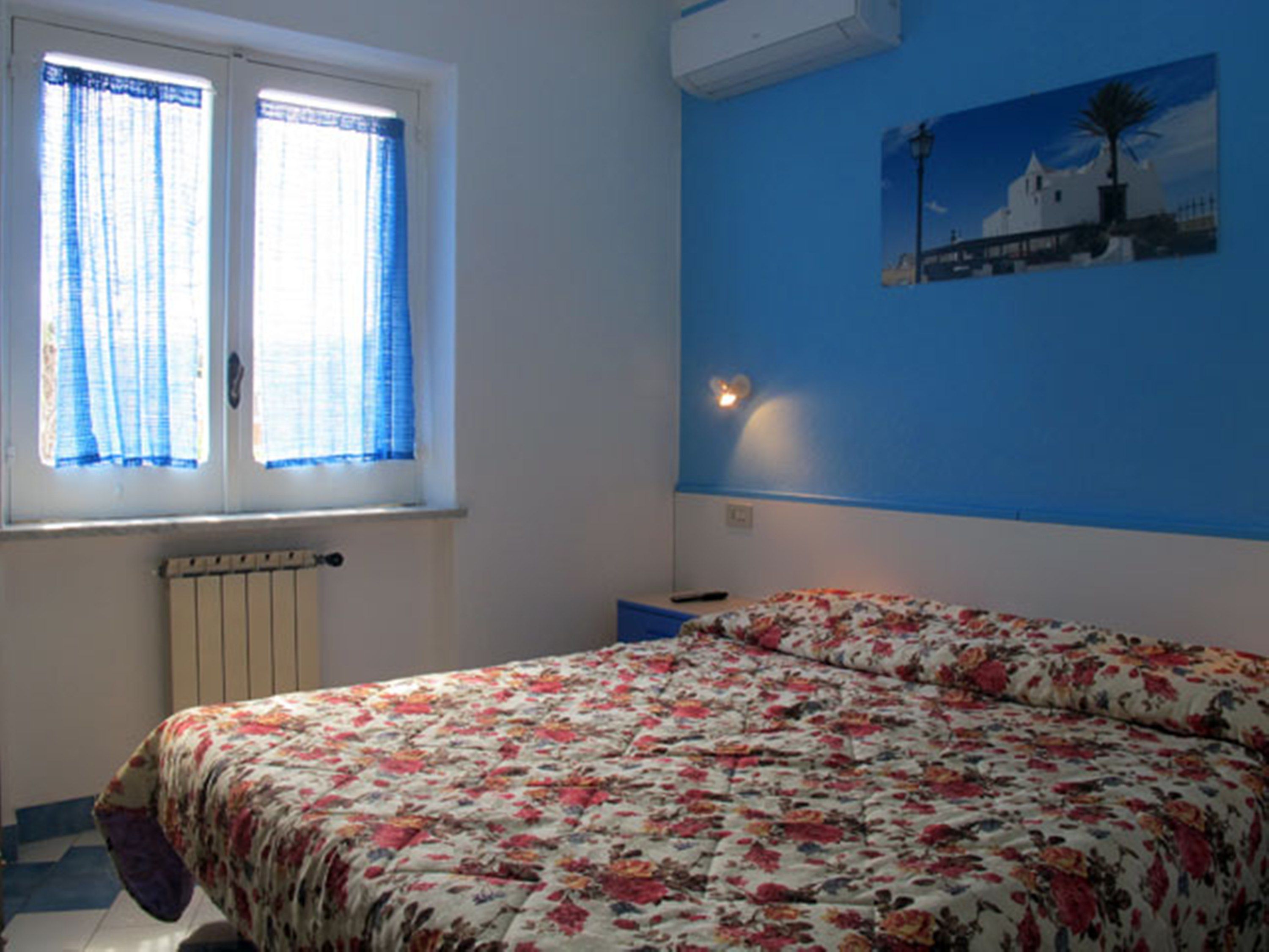Un dormitorio con colcha estampada, ventana con cortinas azules, aire acondicionado de pared y un cuadro de un edificio blanco.
