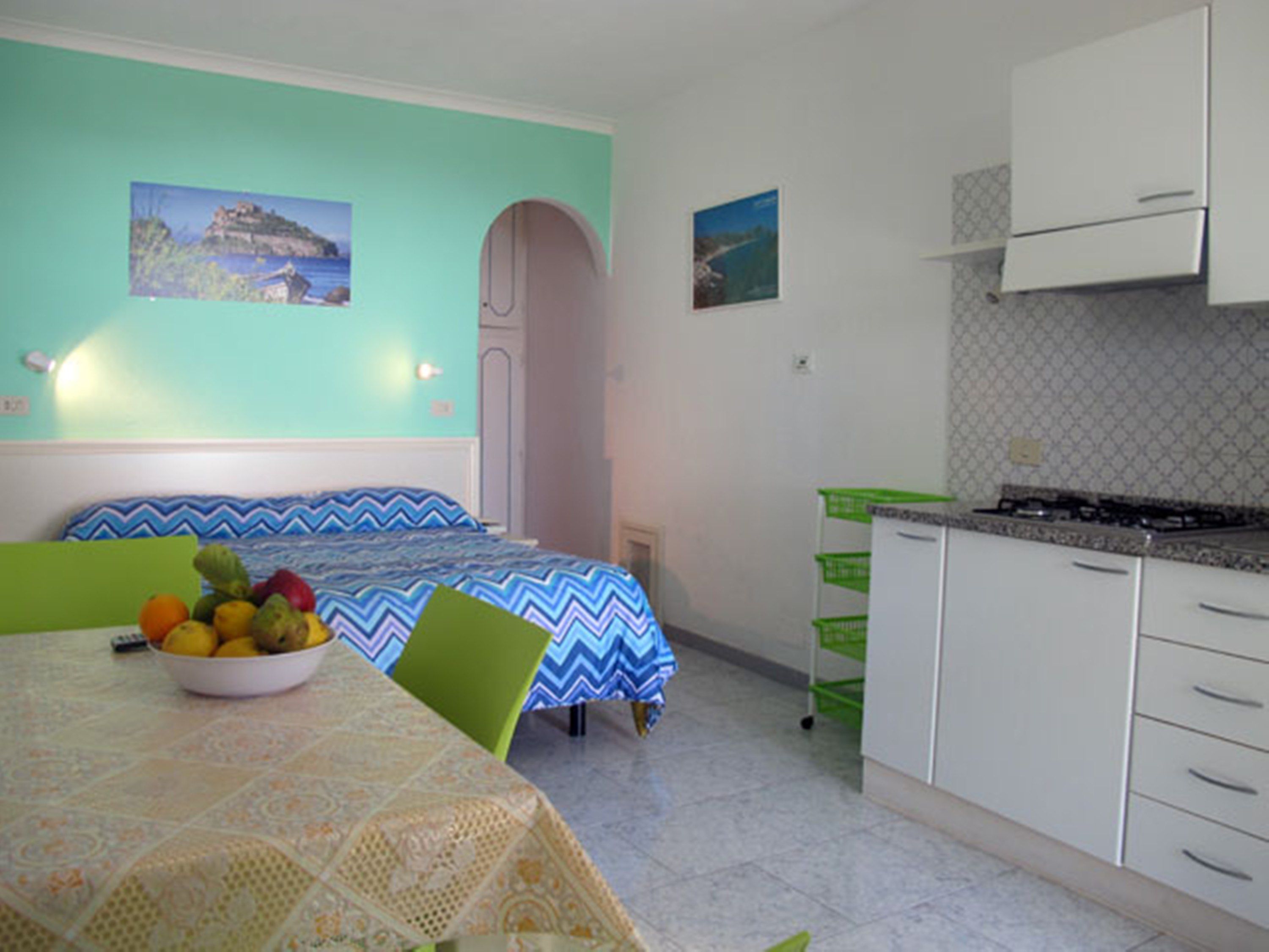 Una habitación pequeña y colorida con una cama, una mesa con un frutero y una kitchenette.