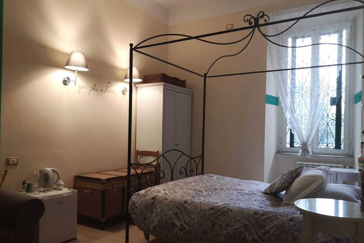 Une chambre à coucher avec un lit à baldaquin, une table de chevet, une chaise, une armoire, un radiateur et une fenêtre avec un rideau.