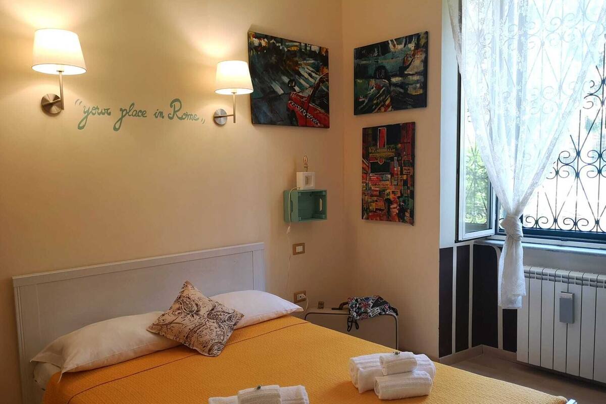 Une chambre d’hôtel avec un lit, deux lampes fixées au mur, une fenêtre avec un rideau et des œuvres d’art accrochées aux murs.