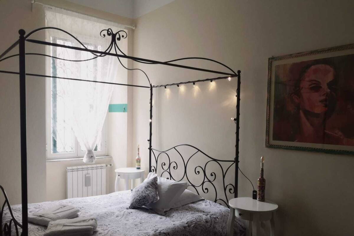 Un lit à baldaquin avec une couvre-lit à motifs blancs et gris, une petite table ronde sur laquelle se trouve une bouteille, et un tableau accroché au mur.