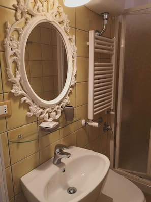 Un baño con un espejo blanco ornamentado, una repisa de vidrio con artículos de aseo, un lavabo blanco y un perchero para toallas.