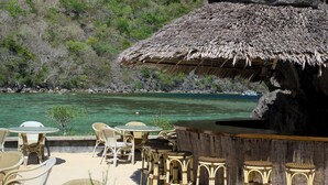A beachside bar with wicker furniture and a thatched roof, overlooking a clear blue sea and a green hillside. - Sangat Island Dive Resort (Coron)
